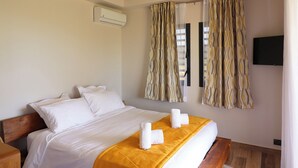 Habitación de hotel con cama, ropa de cama blanca, cabecero de madera, manta amarilla y televisión montada en la pared.