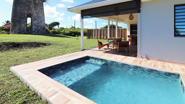 Una piscina con borde de piedra, un porche cubierto con mobiliario exterior y una torre de piedra al fondo.