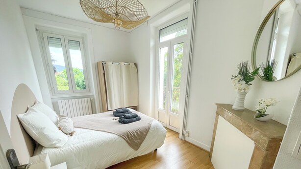 Et soveværelse med seng, natbord og et lille skab. Der er et vindue med gardiner, en dør og en balkon med udsigt over grønne områder.