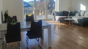 Ein geräumiges Wohnzimmer mit Essbereich, große Schiebeglasfenster, die zu einem Garten führen, und Blick auf Häuser und Hügel.