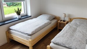 Ein Zimmer mit zwei hölzernen Betten, einem kleinen Nachttisch mit Lampe, einer Zimmerpflanze und einem Fenster mit Jalousien.