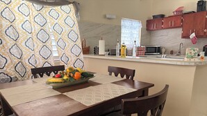 Een eetgedeelte met een houten tafel en stoelen, een fruitschaal, en een keuken op de achtergrond.