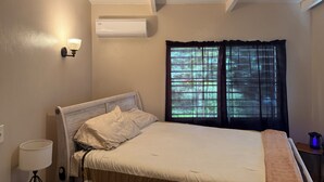 Een slaapkamer met een bed, een wandairconditioner, een raam met gordijnen en een bedlampje.