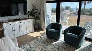 Een woonkamer met een televisie, twee groene fauteuils, een witte bank, een kamerbreed tapijt en uitzicht op het groen.