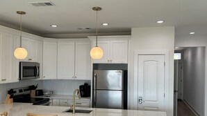 Een moderne keuken met witte kasten, roestvrijstalen apparatuur, een centraal eiland met stoelen, hanglampen en inbouwspots.