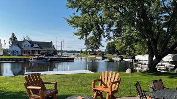 A patio area with wooden Adirondack chairs, a round table, and a view of a calm water body with boats. - Charming 3-bedroom Lake Cottage with Dock, WiFi, & AC on enjoyable Houghton Lake (Houghton Lake)