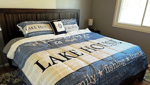 A bedroom with a bed featuring a quilt with 'LAKE HOUSE' and 'family + fishing + friends' text. - Charming 3-bedroom Lake Cottage with Dock, WiFi, & AC on enjoyable Houghton Lake (Houghton Lake)