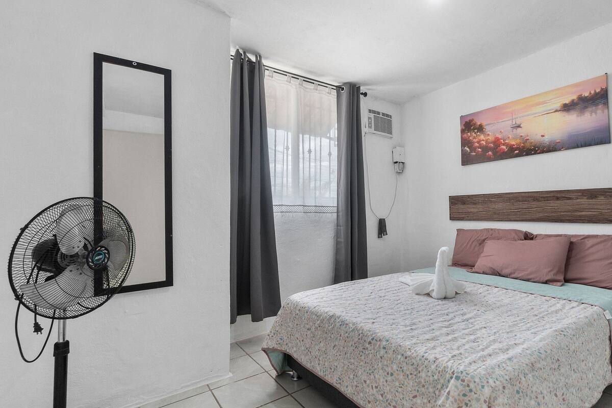 A bedroom with a bed, a fan, a window with curtains, a painting, and a wooden headboard.