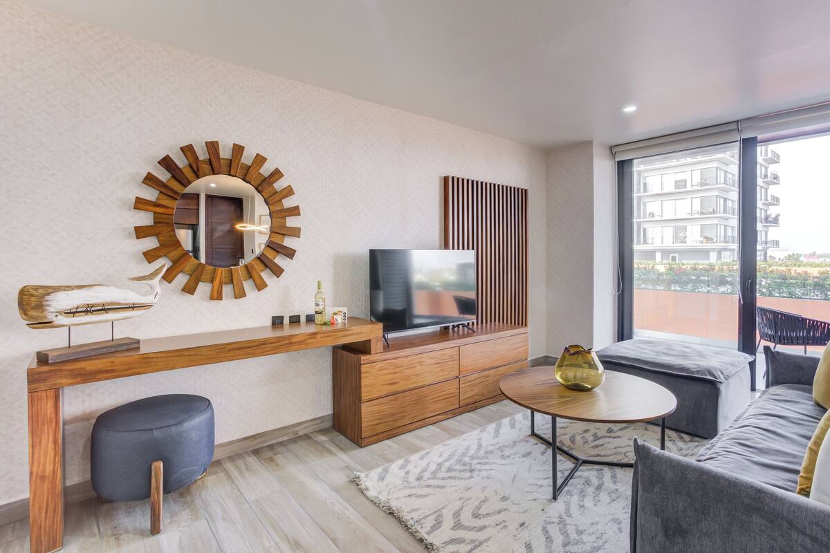 Una sala de estar moderna con televisor, muebles de madera, un espejo redondo y vista al balcón.