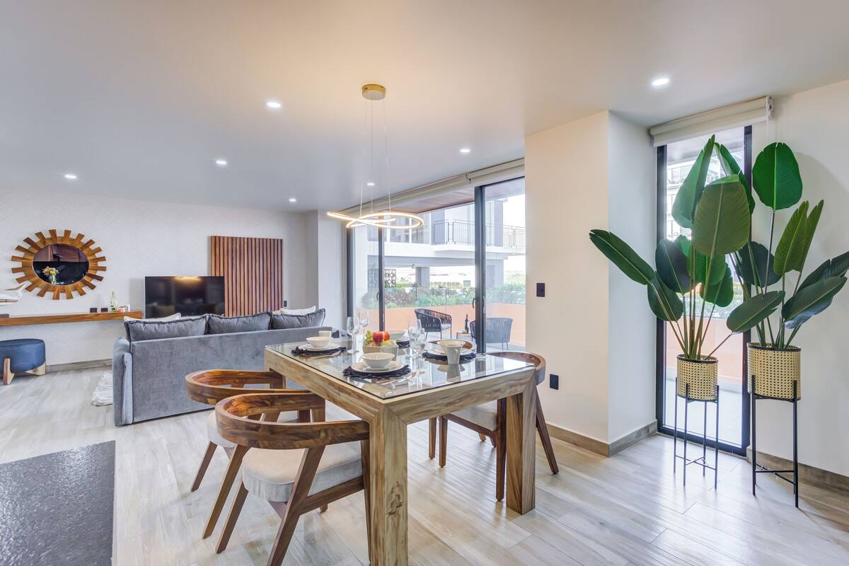 Una sala moderna con una mesa de comedor de madera, un sofá gris y ventanas amplias.