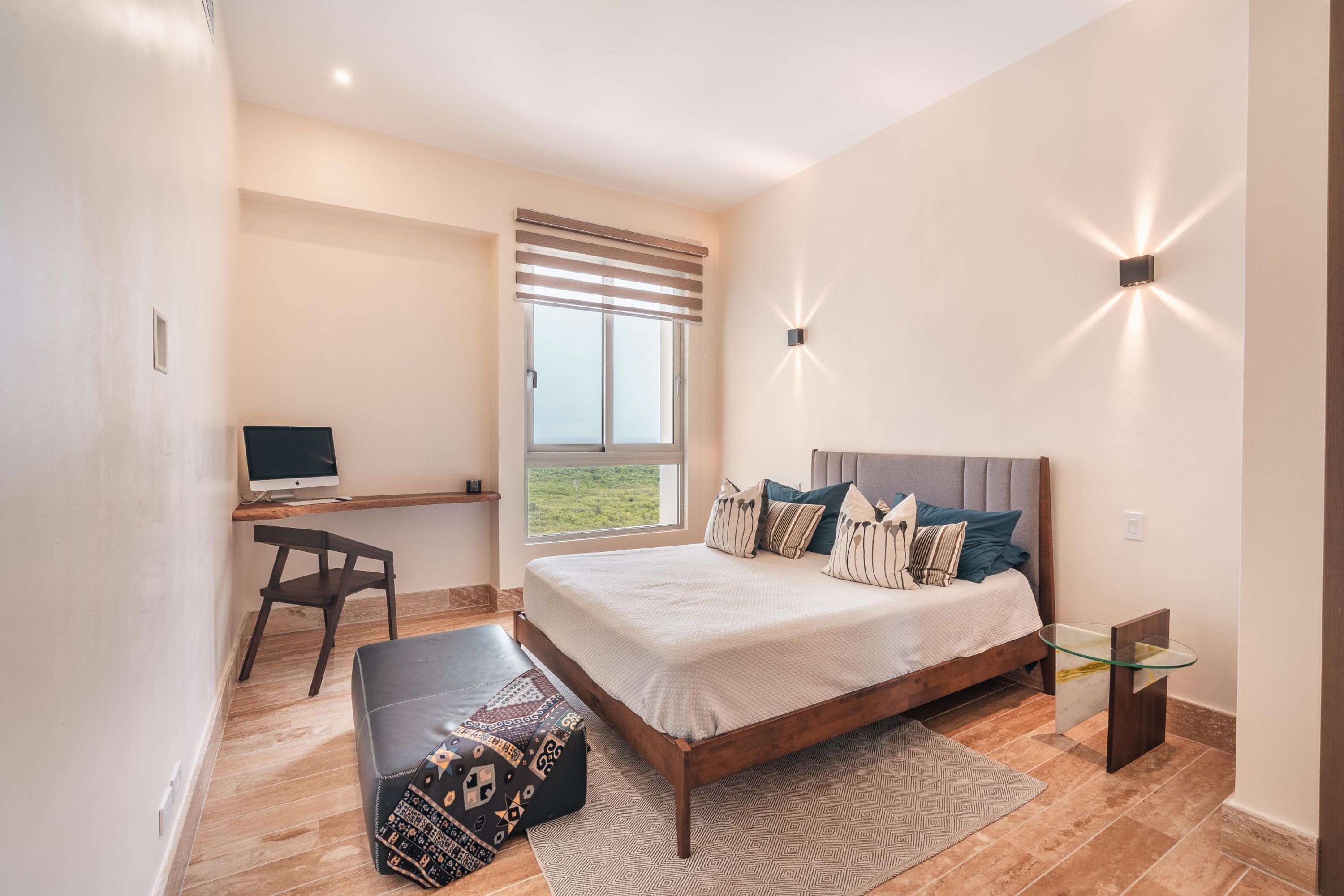 A bedroom with a bed, a desk, a chair, a glass table, and a window with blinds.