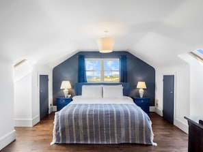 A bedroom with a sloped ceiling, a bed with a striped bedspread, two bedside tables with lamps, and a window with curtains. - Suantraí Cottage (Cashel)