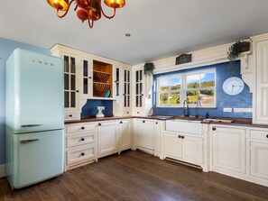 A kitchen with white cabinets, a blue refrigerator, wooden flooring, and a window with a view of greenery. - Suantraí Cottage (Cashel)