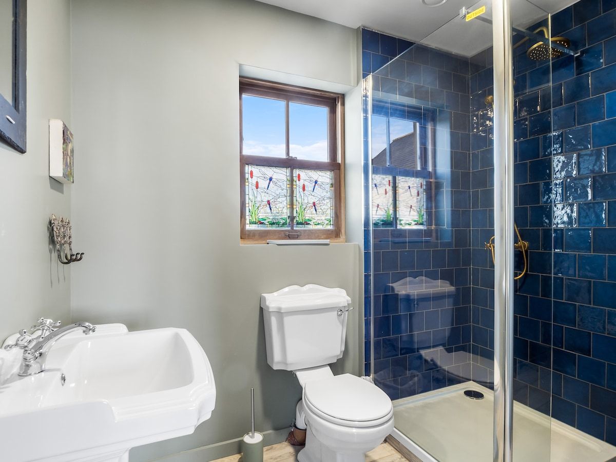 A bathroom with a white toilet, a white sink, a glass shower enclosure, and a window with a view of greenery.