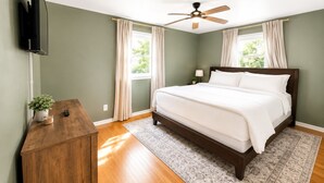A bedroom with a bed, a ceiling fan, a TV, a wooden dresser, and a window with curtains.