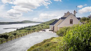 A coastal house with a gravel driveway, a fenced patio, and a view of the sea and hills. - Sky Road Lodge- Beautiful Views (Clifden)