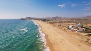Una playa costera con arena, algunos edificios y montañas al fondo.