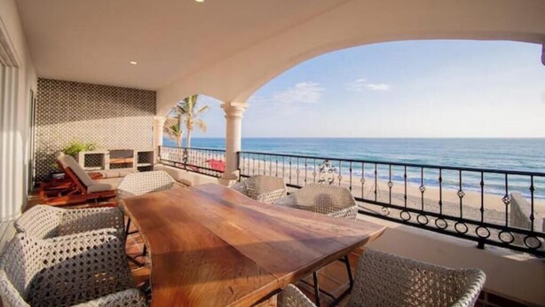 Un espacio de estar techado al aire libre con una mesa de madera y sillas de mimbre, con vista a una playa y una barandilla.
