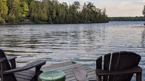 Una terraza de madera con dos sillas Adirondack y una mesa verde, con vistas a un lago tranquilo rodeado de árboles.