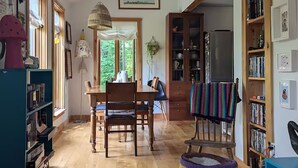 Una sala de estar acogedora con piso de madera, un comedor con mesa y sillas, un sillón colorido y una estantería llena de libros.