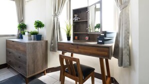 Una habitación con un escritorio de madera, una silla, un aparador y un espejo. Hay plantas sobre el aparador y una ventana con cortinas.