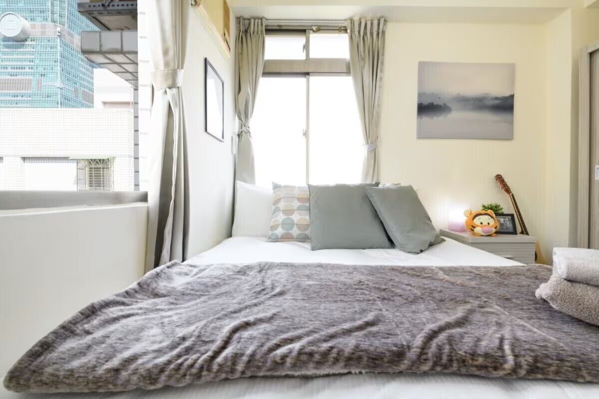 Une chambre à coucher avec un lit, des oreillers, une couverture, une table de chevet avec une lampe, un tableau encadré et une fenêtre avec des rideaux.