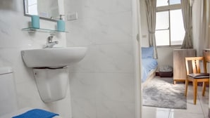 Un baño con un lavamanos blanco, espejo y retrete. Hay una toalla azul sobre el retrete. Se ve una silla de madera y una pequeña mesa en la habitación contigua.