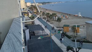 Vista costera desde un balcón con vistas a una playa donde se ven algunas personas caminando, palmeras y un cielo despejado.