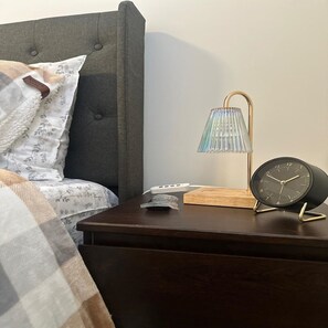 A neatly made bed with a grey headboard, a bedside table with a lamp, a clock, and a remote control. - Renet Suite (Riverview)