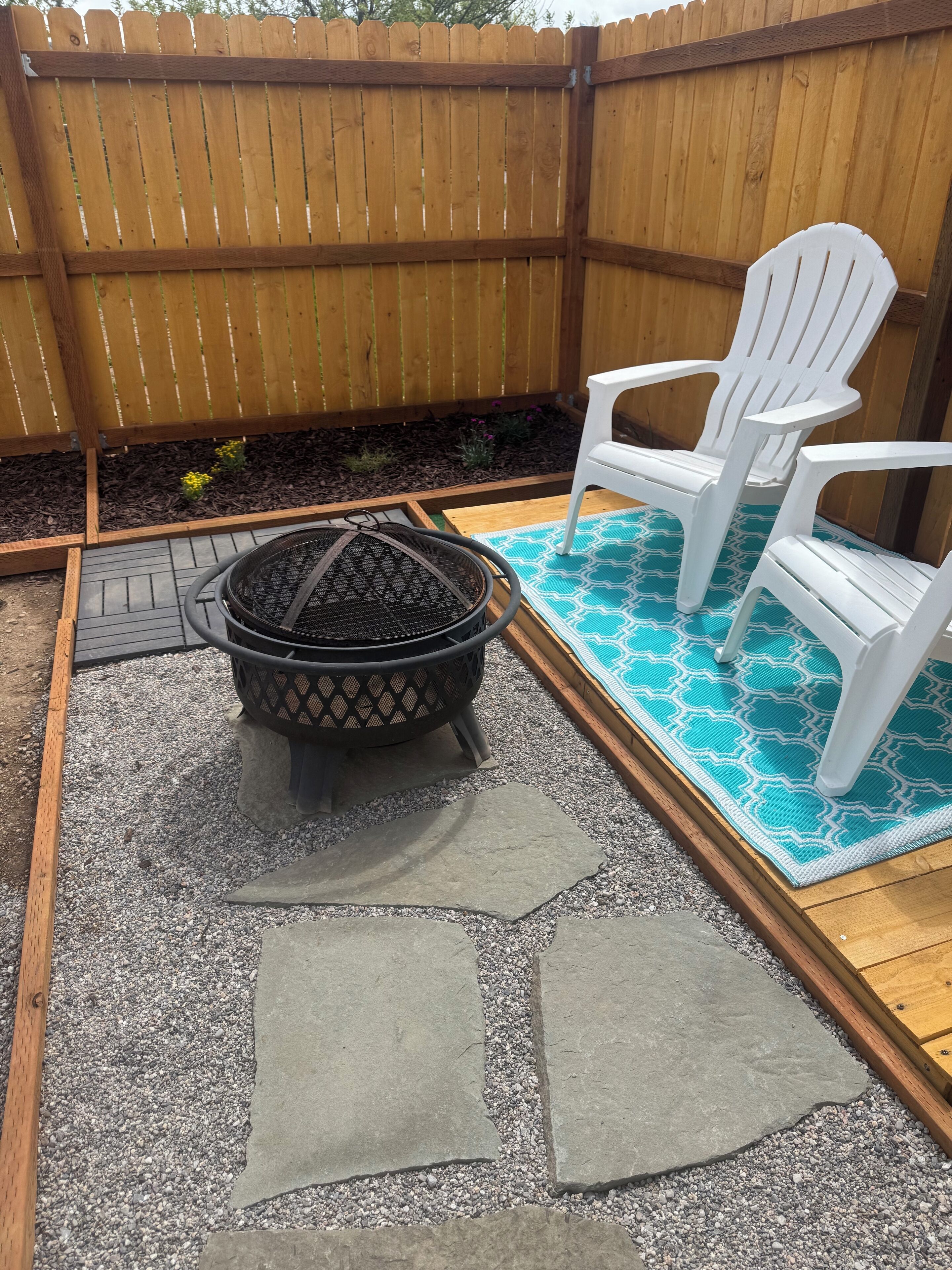 A backyard with a fire pit, two white Adirondack chairs, a blue patterned rug, and a wooden fence.