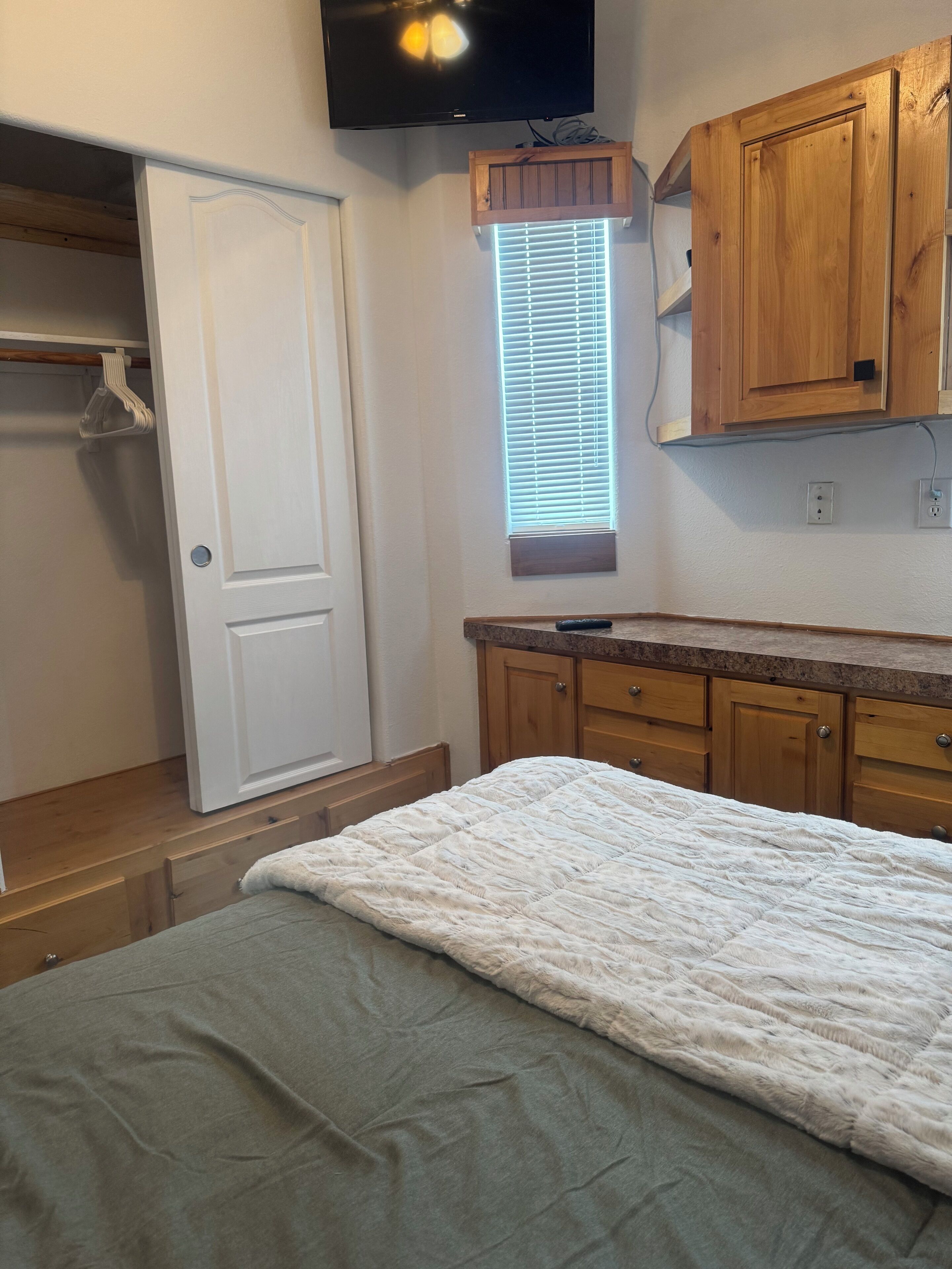A bedroom with a bed, wooden cabinets, a window with blinds, and a television mounted above the bed.