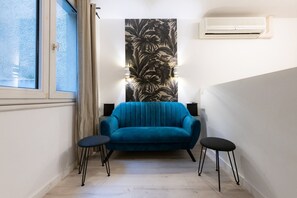 A narrow hallway with a blue sofa, two black stools, a window with curtains, a wall-mounted air conditioner, and a large floral wallpaper. - Part-Dieu  3 - Apartment with AC (Lyon)