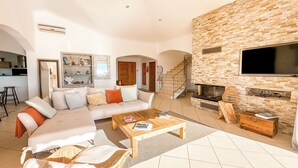 A spacious living room with a large sofa, a coffee table, and a fireplace. There is a staircase leading to an upper level, and a kitchen area is visible in the background. - Villa Golfo Di Sogno (porto vecchio)