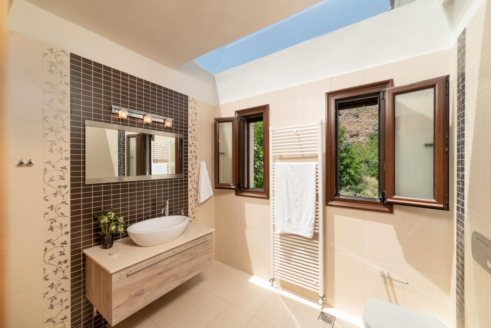 Une salle de bain moderne avec un grand miroir, un lavabo blanc et une fenêtre donnant sur un paysage verdoyant.