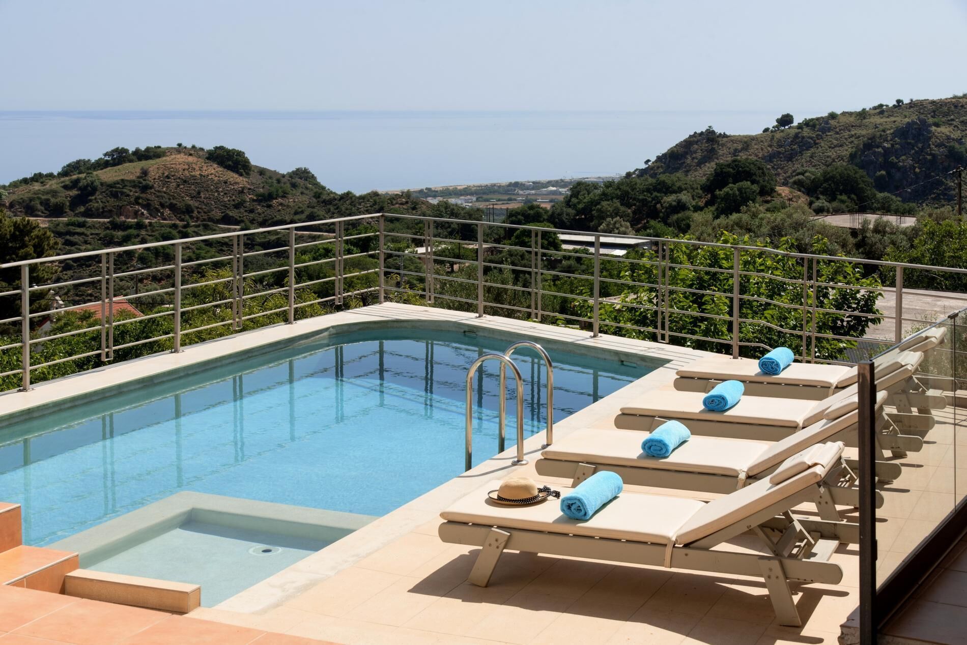 A rooftop pool with clear blue water, surrounded by a metal railing. There are lounge chairs with towels and a hat, and the pool is set against a backdrop of hills and a distant view of the sea.
