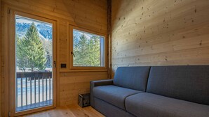 Ein Holzinnenraum mit einem grauen Sofa und zwei Fenstern, die einen Blick auf Bäume und Berge bieten.