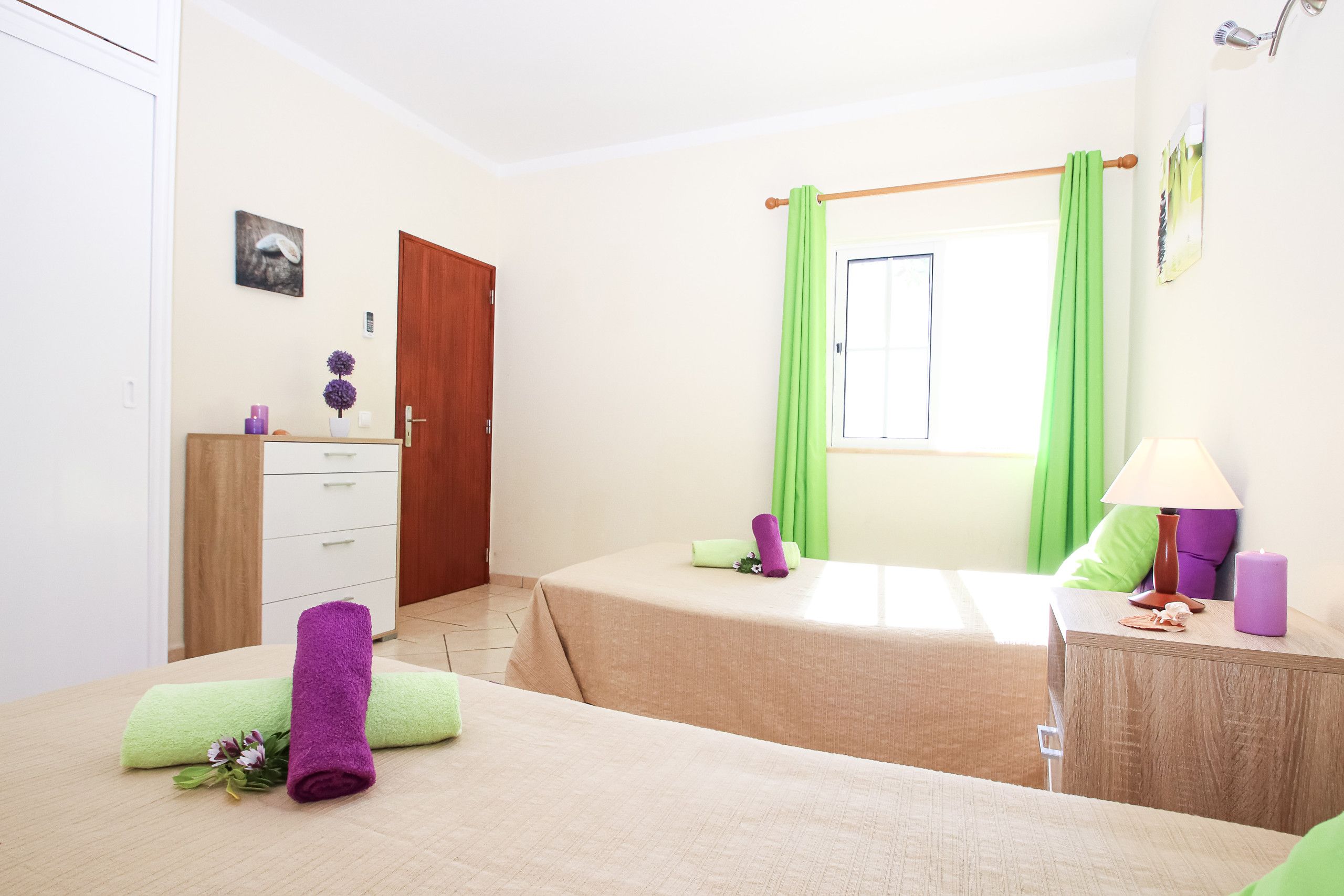 Un dormitorio con cama, cómoda, ventana con cortinas, lámpara y una mesita con velas.