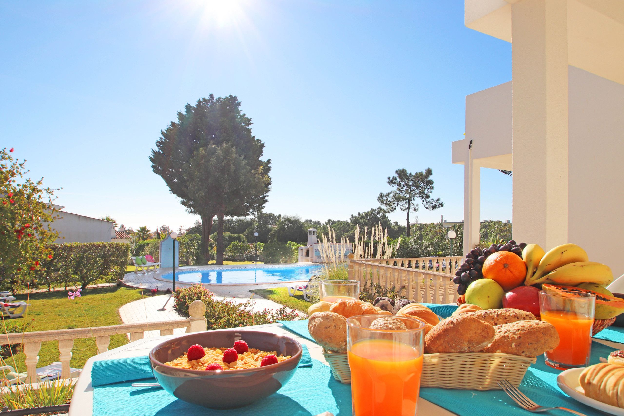 Een ontbijttafel met fruit, gebak en sinaasappelsap op een balkon met uitzicht op een zwembad en tuin.