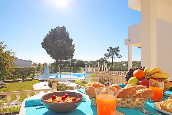 A breakfast table with fruits, pastries, and orange juice on a balcony overlooking a swimming pool and garden.