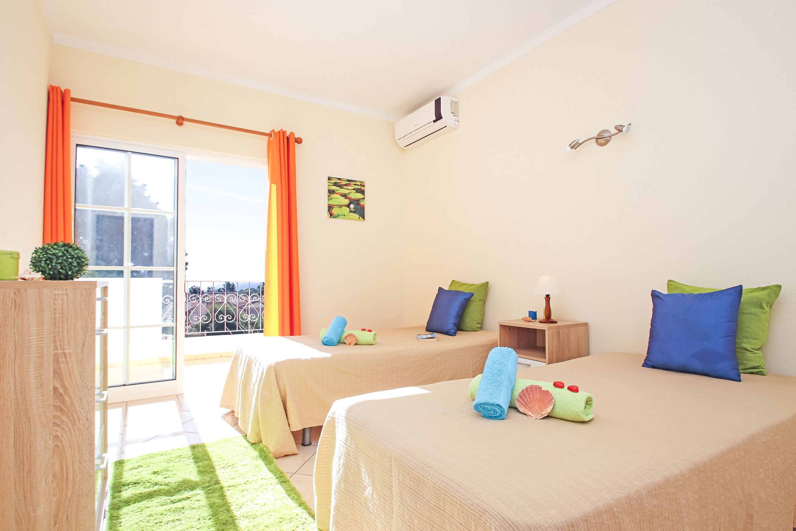 A hotel room with two beds, a balcony, a window with curtains, a wall-mounted air conditioner, and a small table with a lamp.