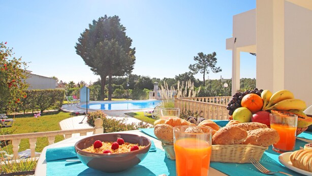 Ein Frühstückstisch mit Früchten, Gebäck und Orangensaft auf einem Balkon mit Blick auf einen Pool und einen Garten.