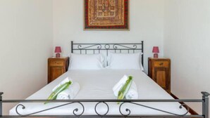 A bed with a metal headboard, two pillows, and two rolled towels with green leaves. There are two bedside tables with lamps and a decorative wall hanging above the bed. - Cozy Apartment in the Heart of Rome- St. Peter's & Vatican (Roma)