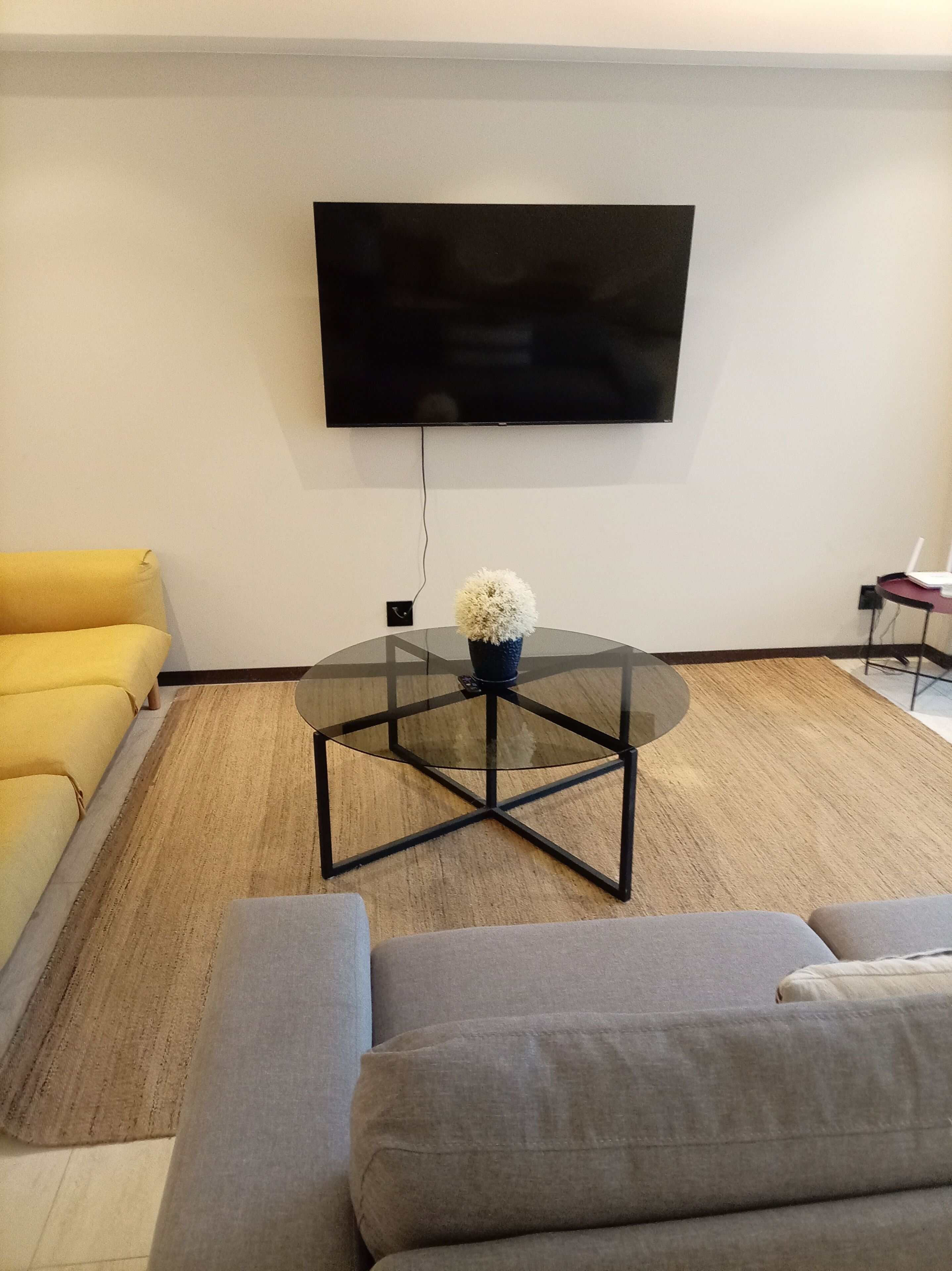 Una sala de estar con una televisión de pantalla plana montada en la pared, una mesa de centro de vidrio con un adorno y dos sofás.