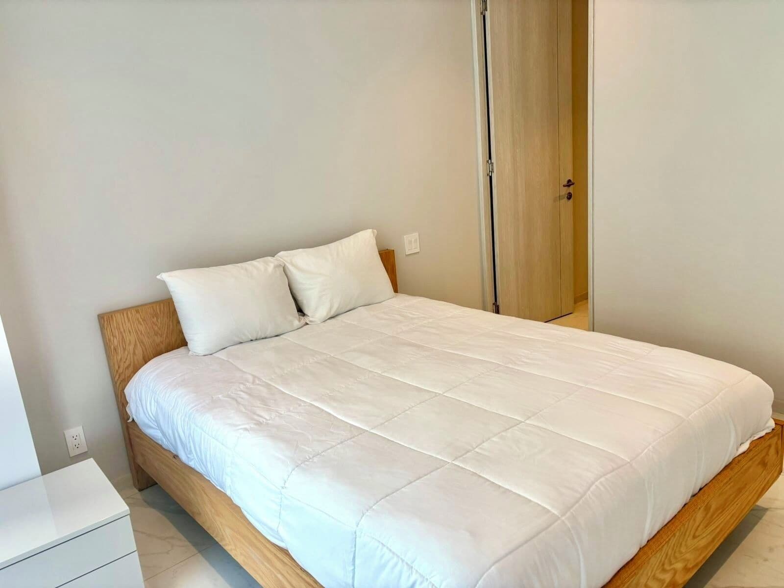 Ein ordentlich bezogenes Bett mit Holz Kopfteil und weißer Bettwäsche in einem schlichten, sauberen Zimmer mit Holztür und einem kleinen weißen Nachttisch.