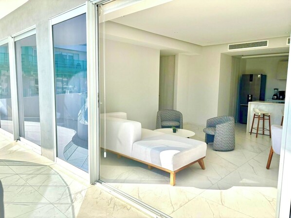 Una sala de estar moderna con puerta de vidrio que da a un balcón, una pequeña zona de cocina y un espacio de estar con sofá y sillones.