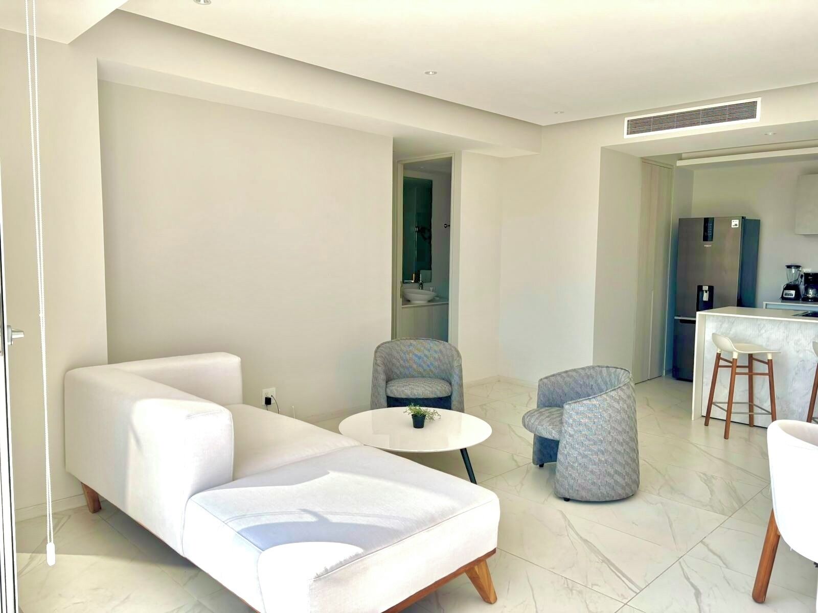 A modern living room with a white sofa, two patterned armchairs, a small round table, and a kitchen area with a refrigerator and bar stools.