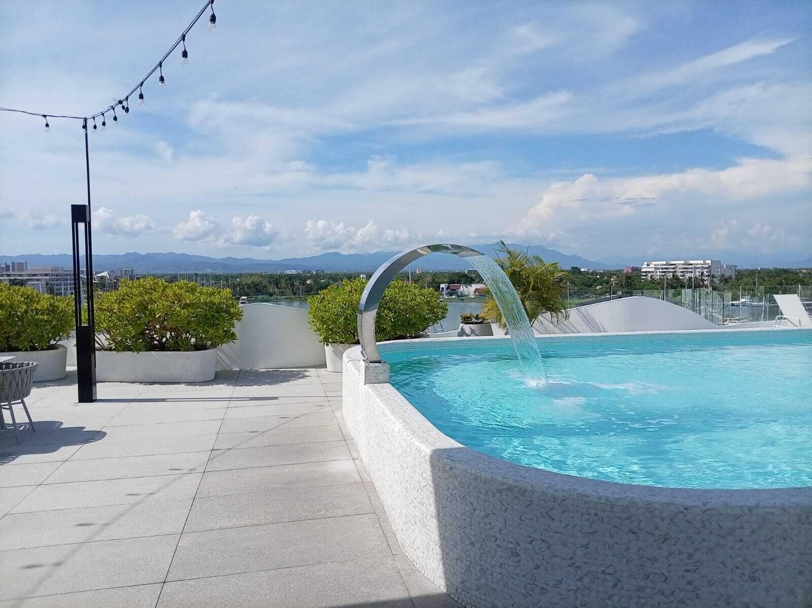 A rooftop pool with a waterfall feature, surrounded by potted plants and a clear view of distant mountains.