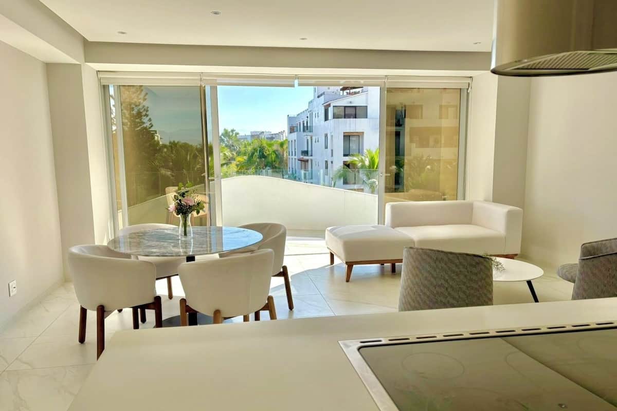 Una sala de estar moderna con un ventanal que da a un balcón, muebles blancos y vistas a edificios y zonas verdes.