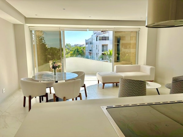 Un salon moderne doté d’une porte vitrée donnant sur un balcon, de meubles blancs et offrant une vue sur des immeubles et de la verdure.