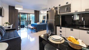 A modern hotel room with a sofa, dining table, and a kitchen area with a refrigerator, microwave, and a small sink. - 112 Rosemari's Apartments Studio Premium Centro SP Garage & Pool & Gym (República São Paulo)